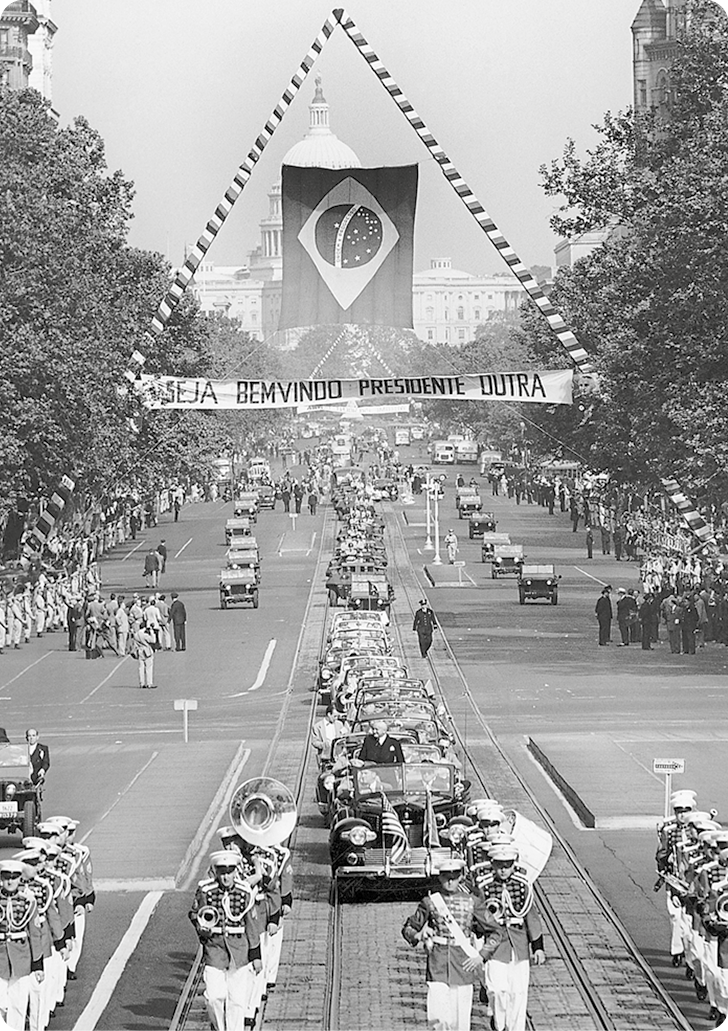 Fotografia em preto e branco. Um desfile, com carros e homens uniformizados tocando instrumentos de sopro e percussão. No segundo plano, a bandeira do Brasil dentro de um triângulo. Abaixo da bandeira, uma faixa com o texto: SEJA BEM VINDO PRESIDENTE DUTRA.