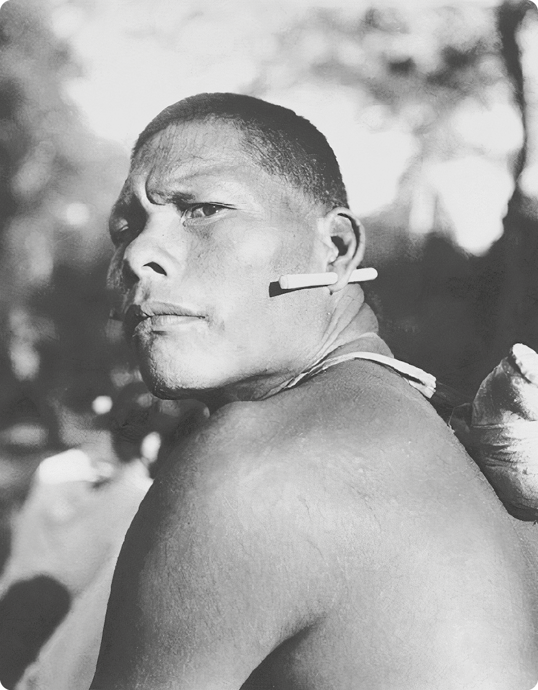 Fotografia em preto e branco. Destacando o busto de um homem indígena com um alargador na orelha.