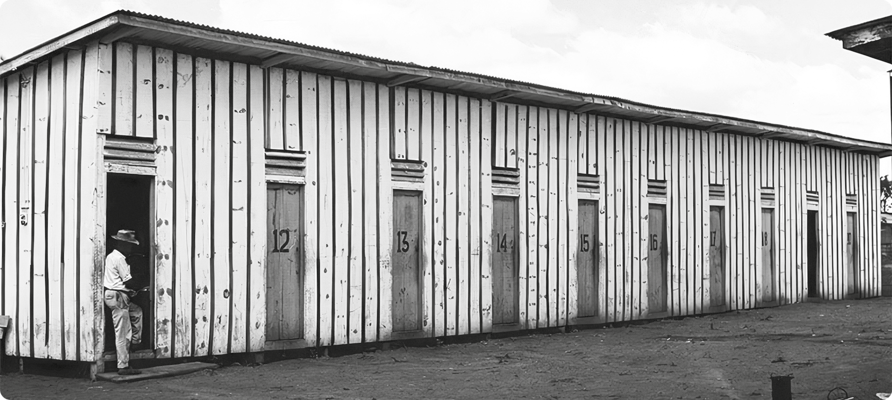 Fotografia em preto e branco. Uma construção de madeira com muitas portas. Um homem usando chapéu está de pé em uma das portas.