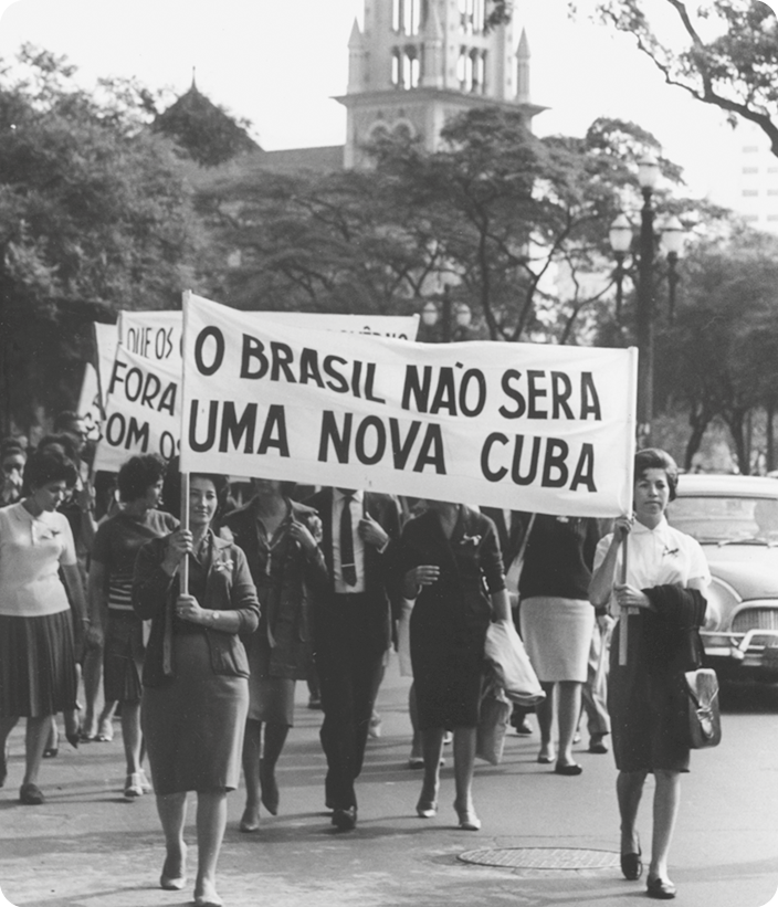 Fotografia em preto e branco. Mulheres caminhando em uma rua, segurando um cartaz com o texto: O BRASIL NÃO SERÁ UMA NOVA CUBA.