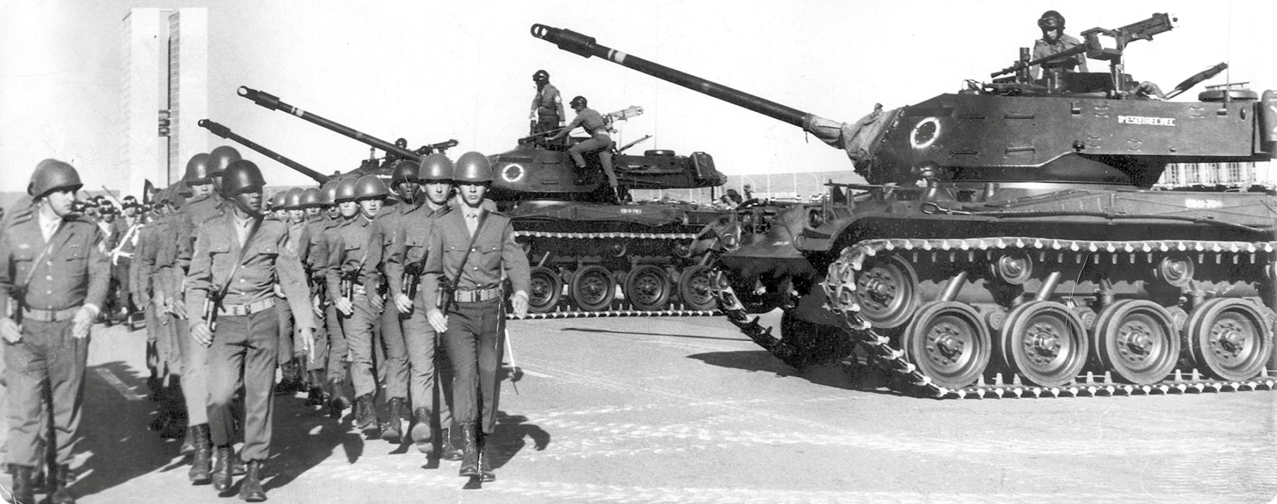 Fotografia em preto e branco. À esquerda, soldados em fileiras caminhando. À direita, tanques de guerra em uma rua.