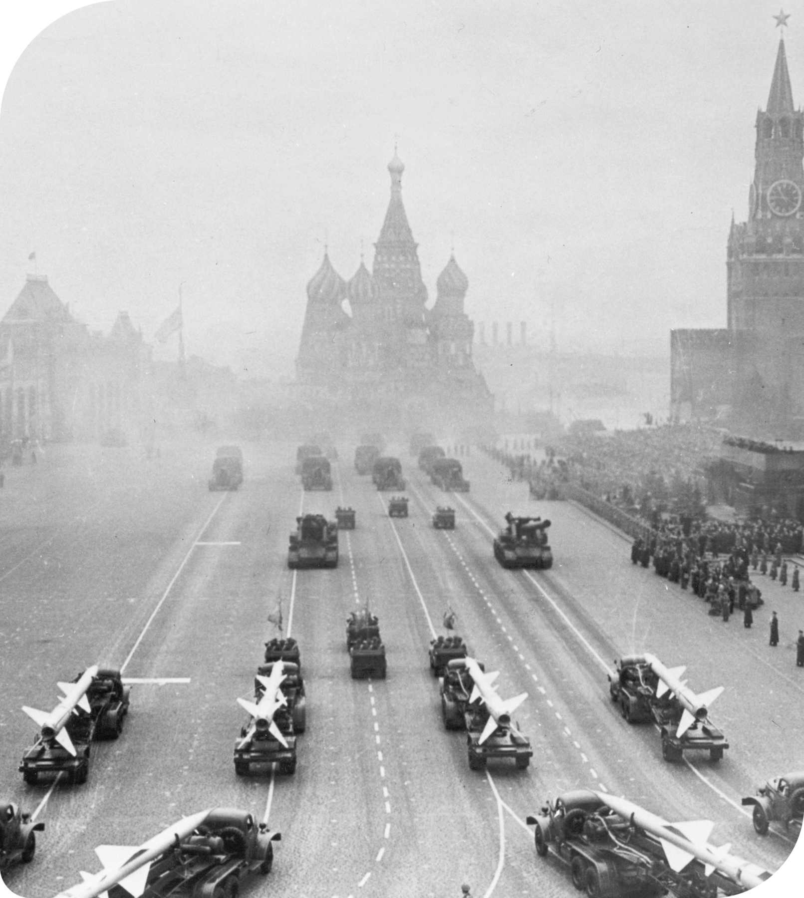Fotografia em preto e branco. Vista de uma avenida. Nela, há vários caminhões carregando mísseis e tanques de guerra. Ao fundo, a silhueta de um castelo com várias torres arredondadas. À direita, várias pessoas assistindo ao desfile.