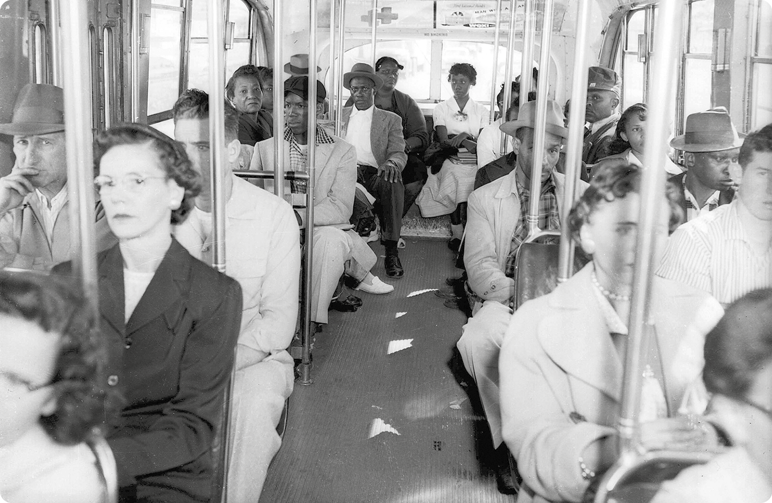 Fotografia em preto e branco. Interior de um ônibus. Na parte da frente, mulheres e homens brancos sentados. Na parte traseira, mulheres e homens negros sentados.