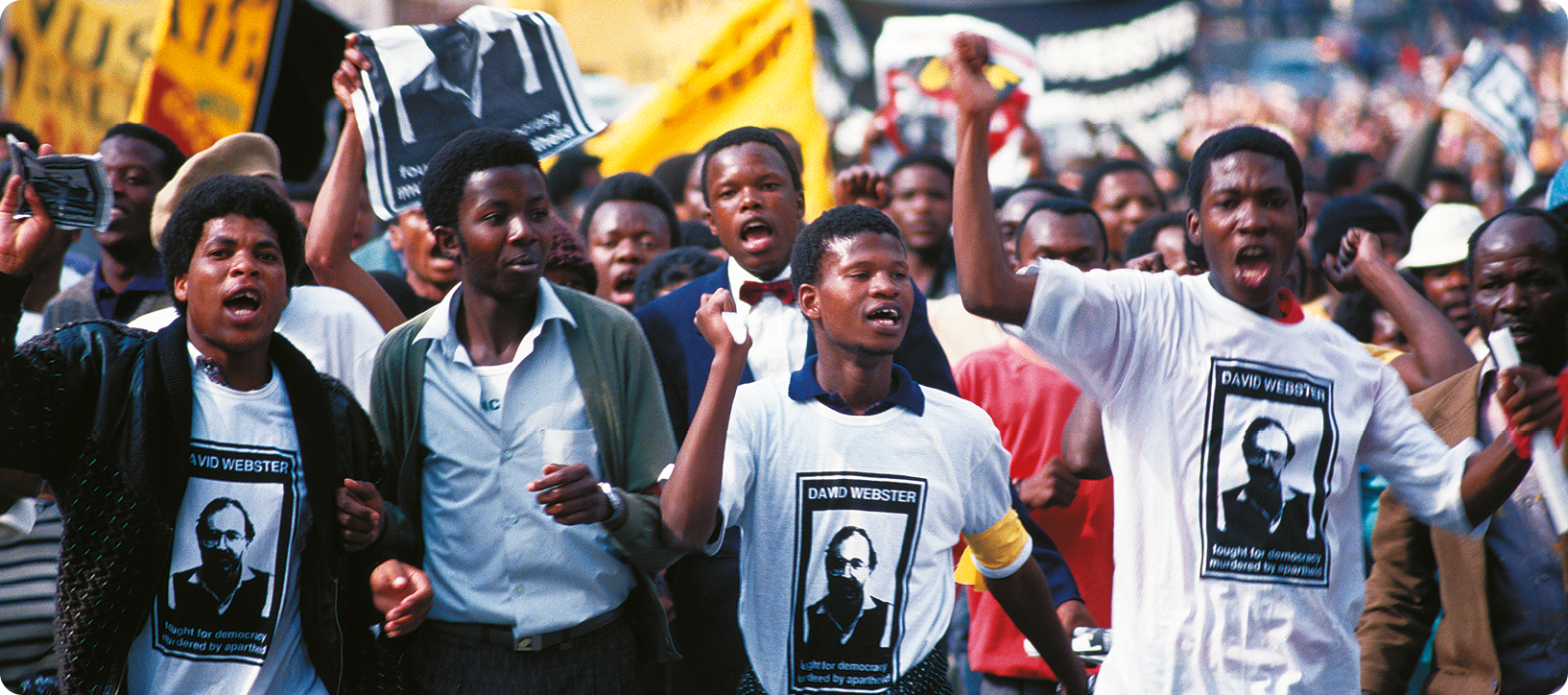Fotografia. No primeiro plano, jovens negros usando uma camisa com a fotografia de um homem. Ao fundo, pessoas estão em uma rua carregando cartazes e faixas.