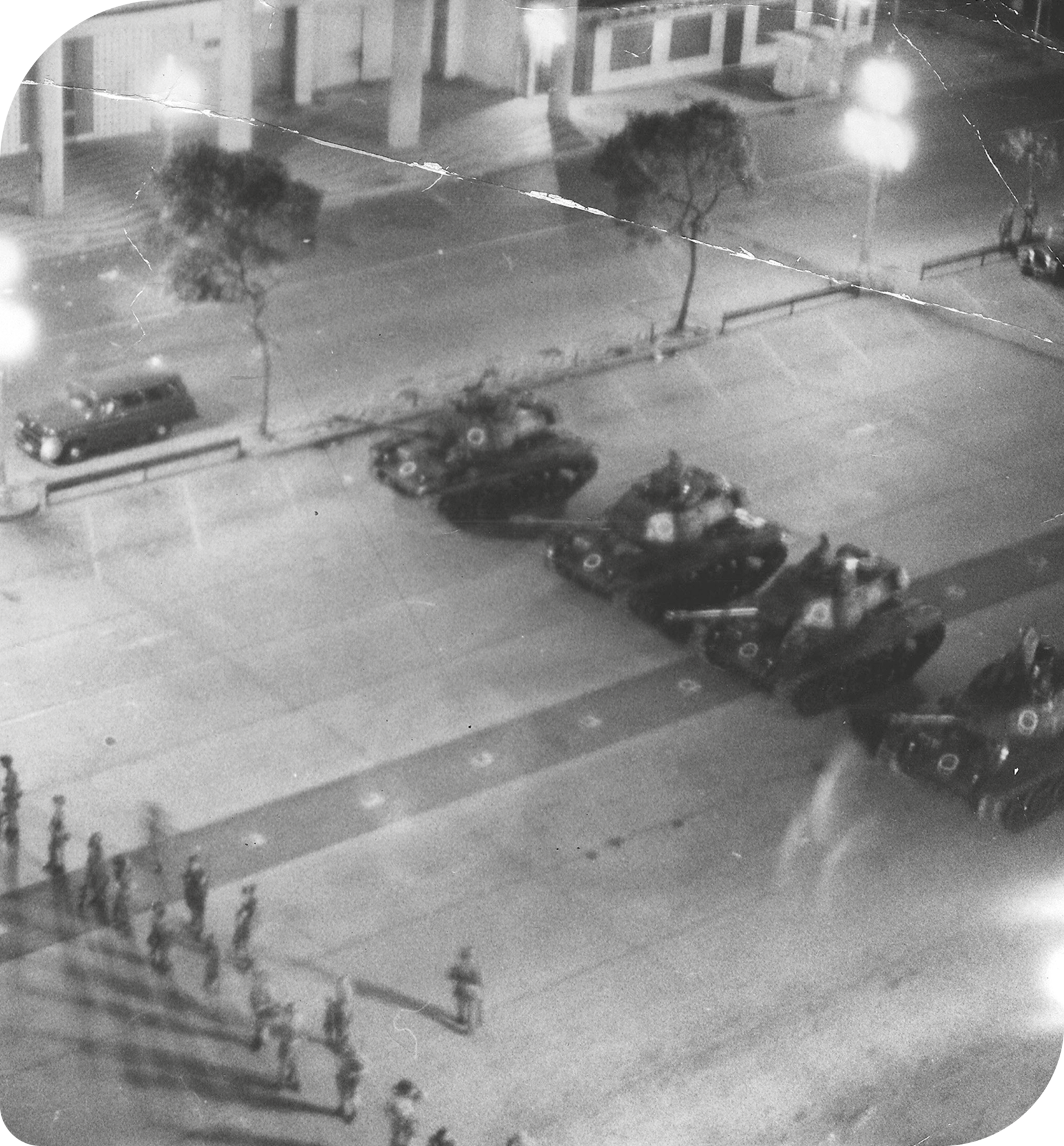 Fotografia em preto e branco. Vista aérea de uma avenida. À esquerda, várias pessoas enfileiradas. À direita, quatro tanques de guerra.