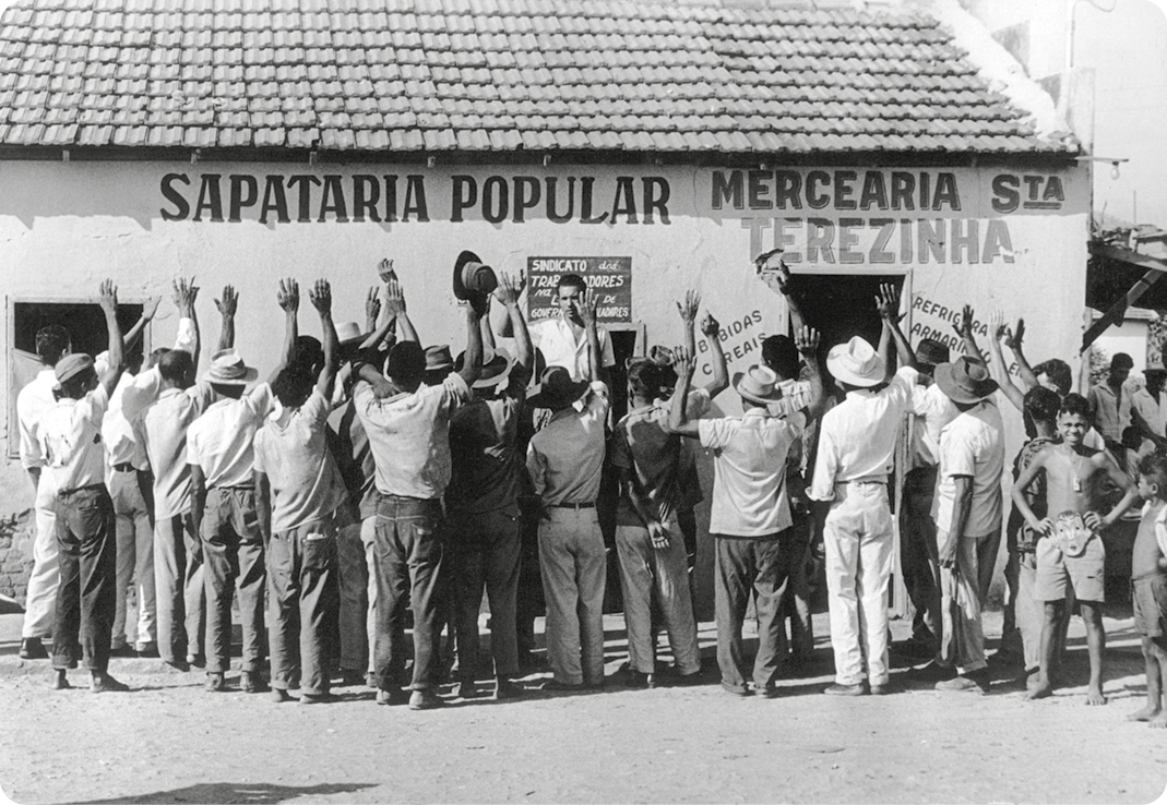 Fotografia em preto e branco. No primeiro plano, vários homens de costas, segurando chapéu com as mãos. Eles estão olhando na direção de uma construção com o texto: SAPATARIA POPULAR e MERCEARIA STA. TEREZINHA.