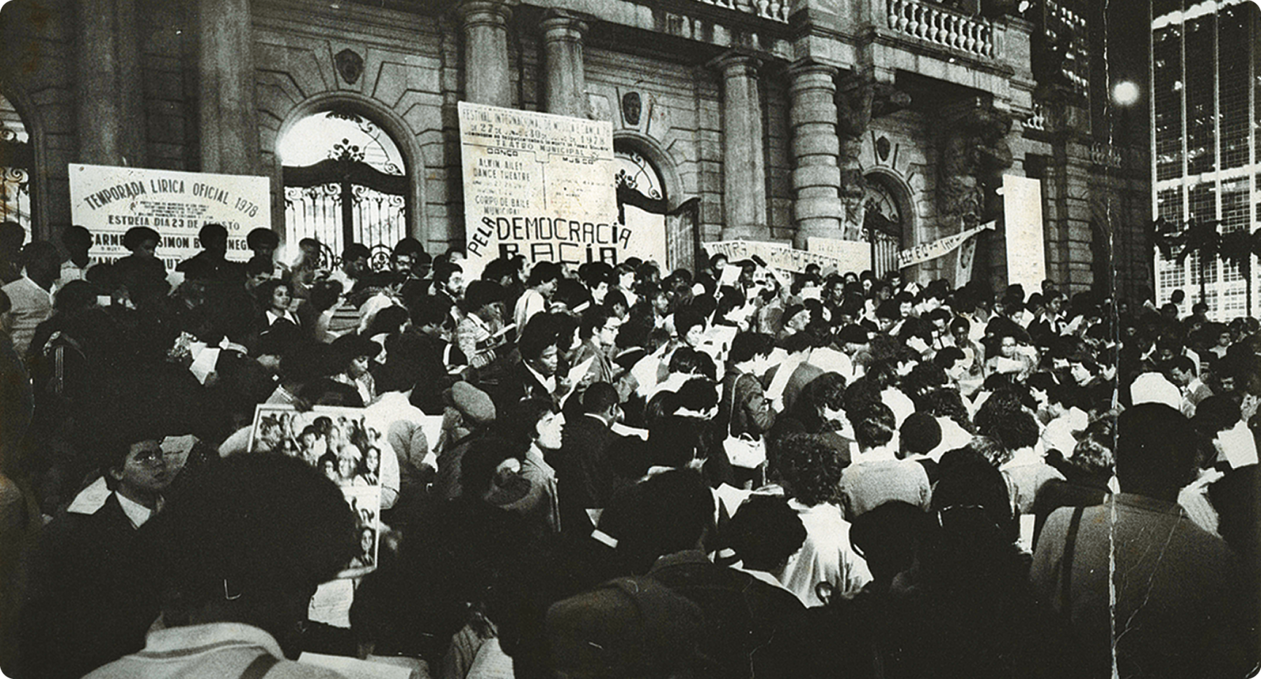 Fotografia em preto e branco. Diversas pessoas em uma rua segurando cartazes e placas.