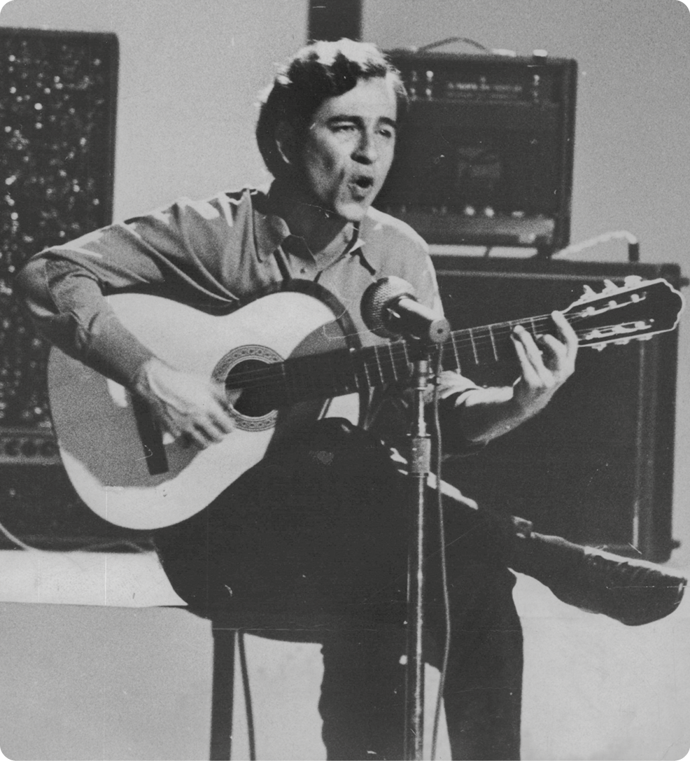 Fotografia em preto e branco. Um homem com cabelos ondulados, usando camisa e calça. Ele está sentado, segurando com as mãos um violão e a boca perto de um microfone.