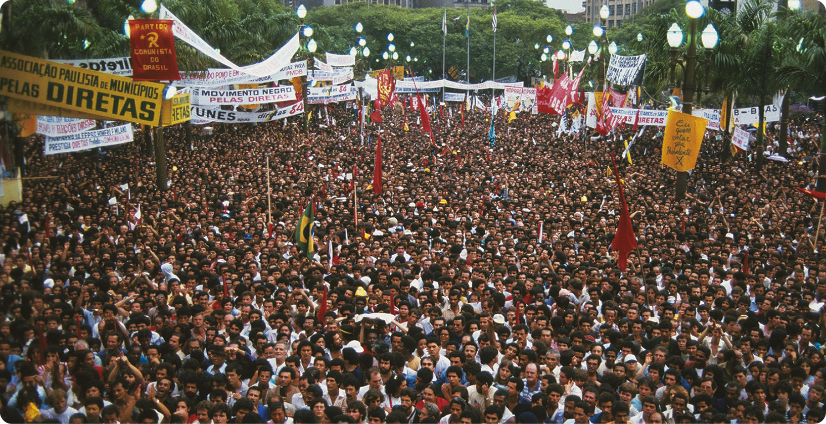Fotografia. Muitas pessoas segurando cartazes com o texto: DIRETAS, e bandeiras vermelhas e do Brasil..