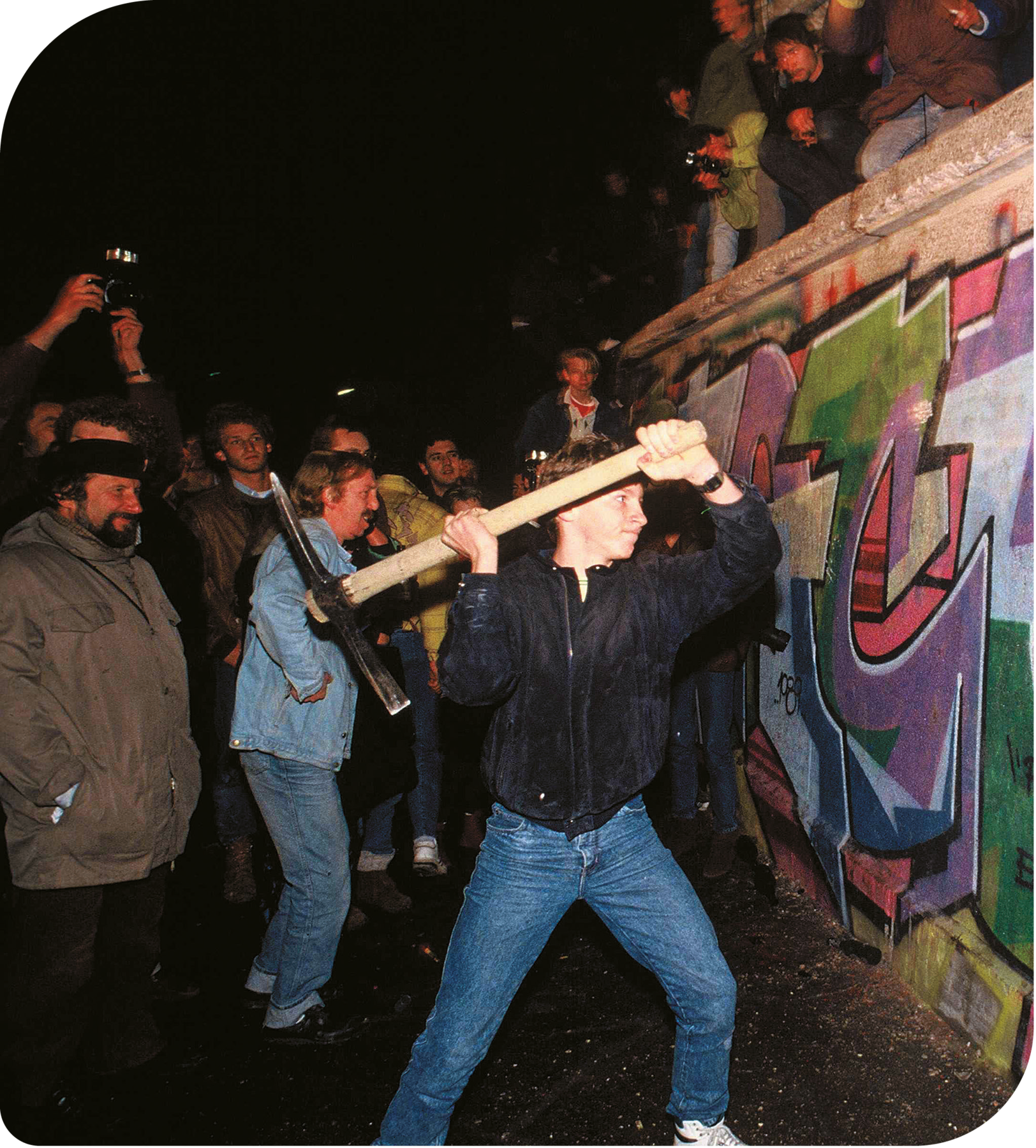 Fotografia. Um jovem, usando casaco e calça jeans, está de perfil, segurando com as mãos uma picareta na direção de um muro. Ao redor, diversas pessoas olhando.