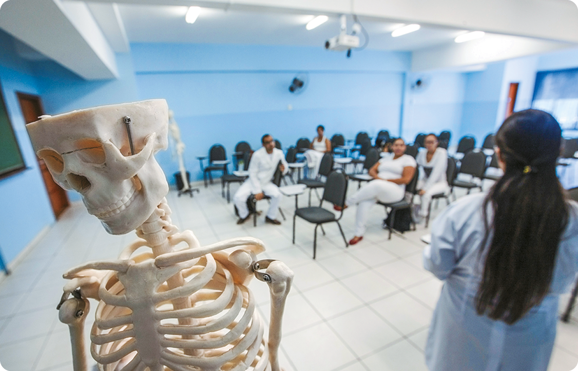 Fotografia. Vista de uma sala de aula. Em primeiro plano, um esqueleto. Ao lado, pessoas usando jaleco, uma está em pé e as outras sentadas em carteiras.