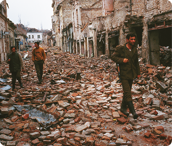 Fotografia. Três pessoas caminhando sobre escombros de um edifício.