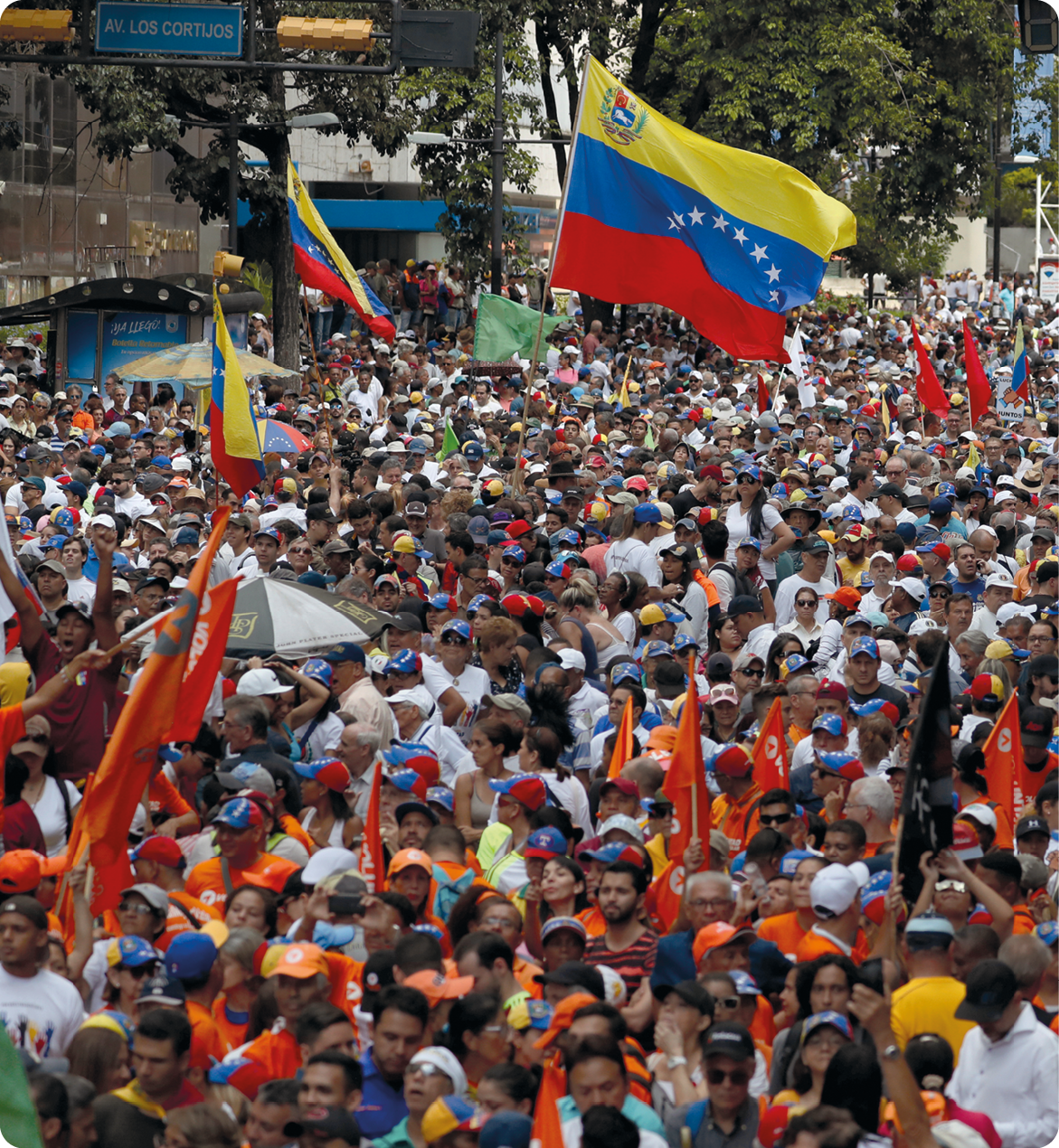 Fotografia. Vista aérea. Várias pessoas vestindo boné cor laranja. Ao centro, algumas bandeiras da Venezuela.