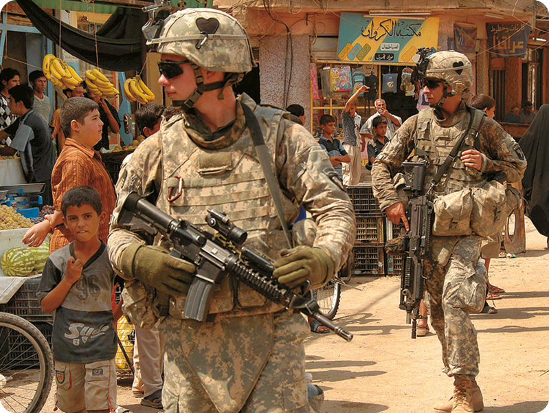Fotografia. Soldados usando fardas militares e segurando armas com as mãos. Eles estão caminhando em uma rua de terra. Ao redor, pessoas caminhando.