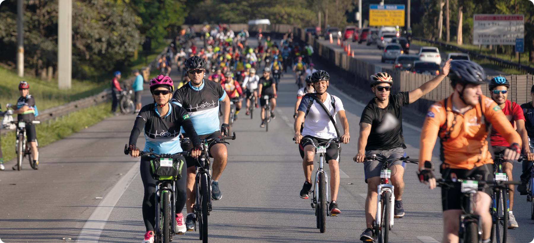 Fotografia. Diversas pessoas usando capacete e roupas coloridas andando de bicicleta em uma rua.