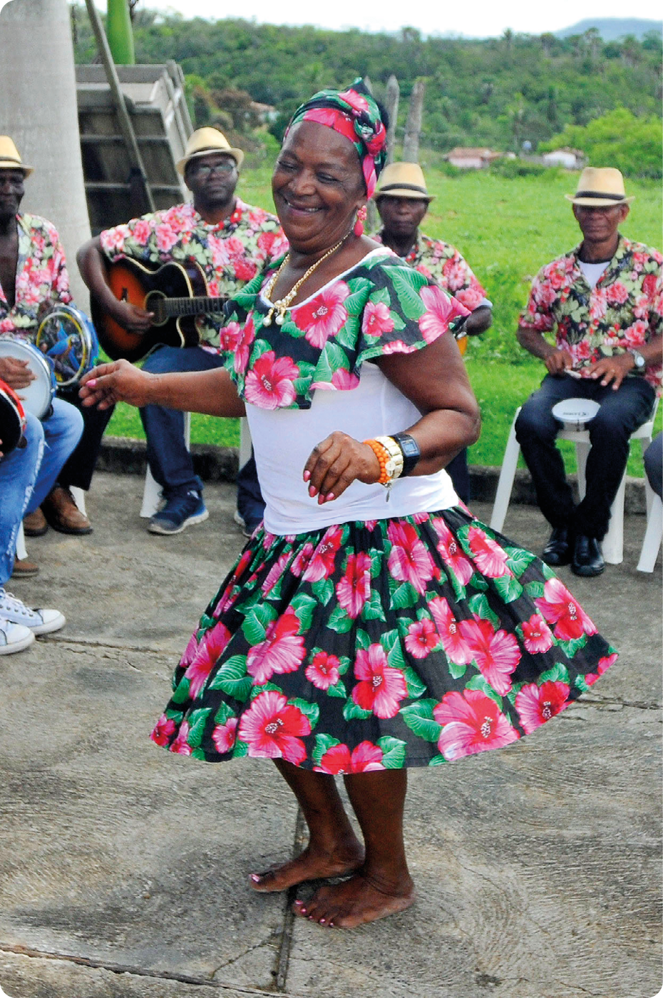 Fotografia. Uma mulher negra, usando lenço florido na cabeça, vestido rodado com babado. Ela está descalça e dançando. Atrás, homens negros usando chapéu, camisa florida e calça. Eles estão sentados, segurando com as mãos instrumentos musicais.