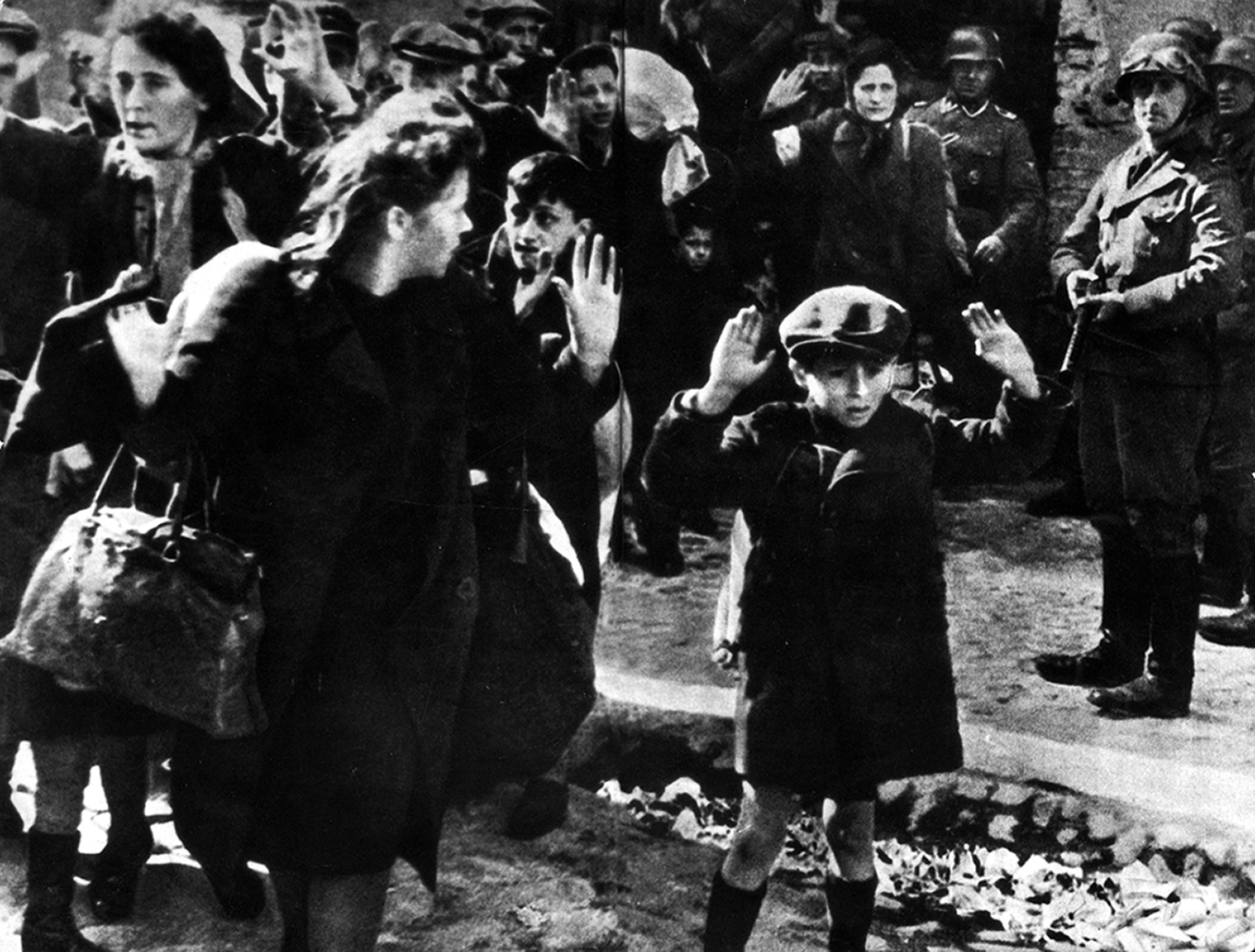 Fotografia em preto e branco. Mulheres e crianças com as mãos para cima em uma rua com soldados apontando armas para elas.