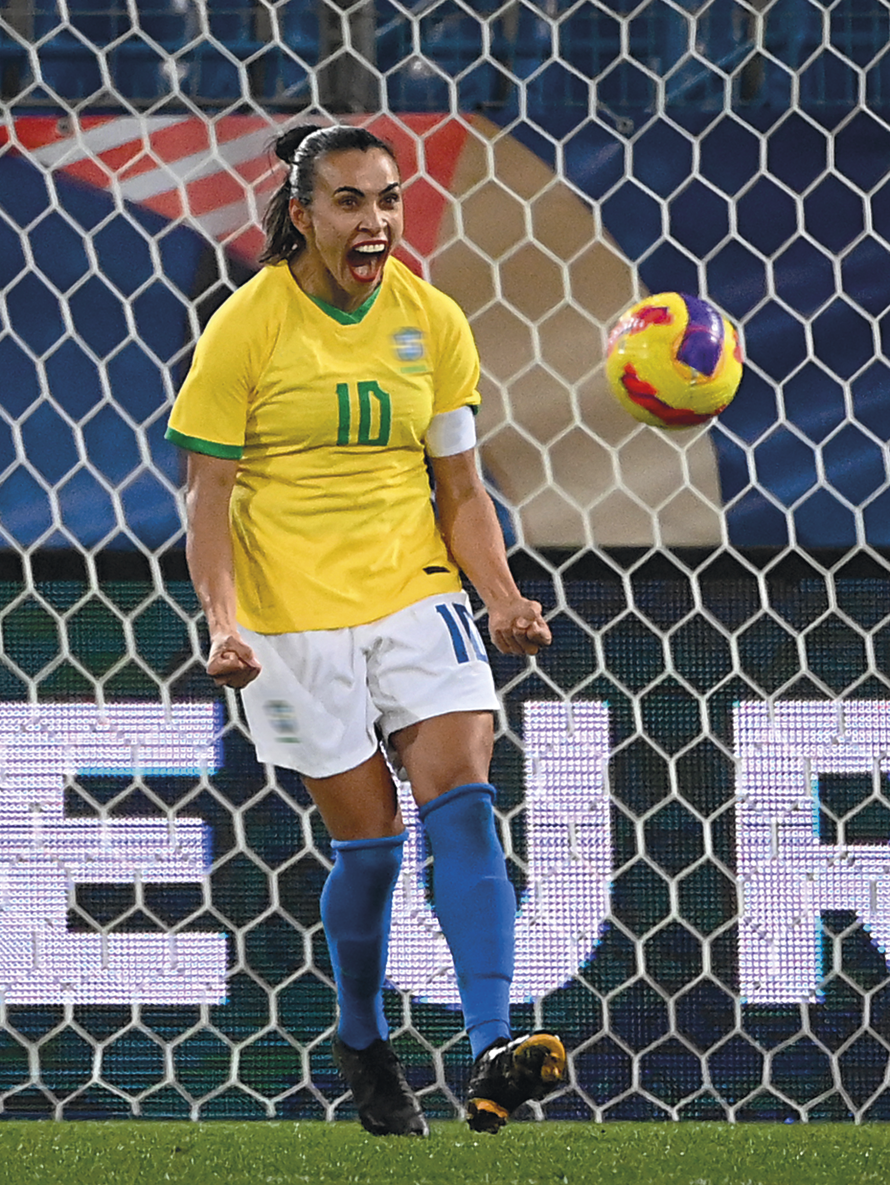 Fotografia. Mulher com cabelos presos, usando uniforme da seleção brasileira de futebol. Ela está com a boca aberta. Atrás, uma bola e uma rede.