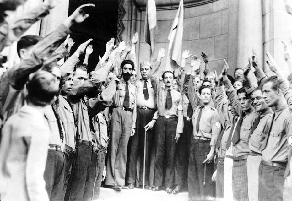 Fotografia em preto e branco. Diversos homens, usando camisas, calças e gravatas, estão com o braço direito levantado na diagonal.