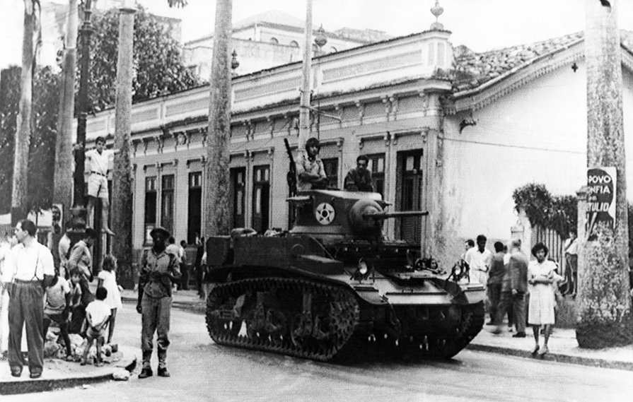 Fotografia em preto e branco. Um tanque de guerra em uma rua. Há dois soldados sobre ele e outro em pé, na lateral do tanque. Atrás, um edifício. Ao redor, pessoas caminhando.