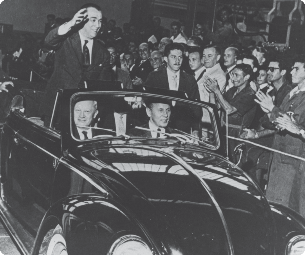Fotografia em preto e branco. À esquerda, homem usando terno. Ele está de pé e dentro de um carro aberto, acenando com uma das mãos. Nas laterais, pessoas em pé batendo palmas.