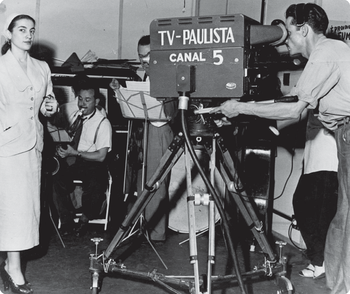 Fotografia em preto e branco. À esquerda, uma mulher usando terno e saia. Ao lado, um homem segurando uma câmera filmadora com a placa: TV-PAULISTA CANAL 5. Ao fundo, homens tocando instrumentos musicais.