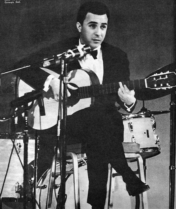 Fotografia em preto e branco. Homem com cabelos pretos usando um terno com gravata borboleta. Ele está sentado, segurando um violão com as mãos e a boca próxima ao microfone.