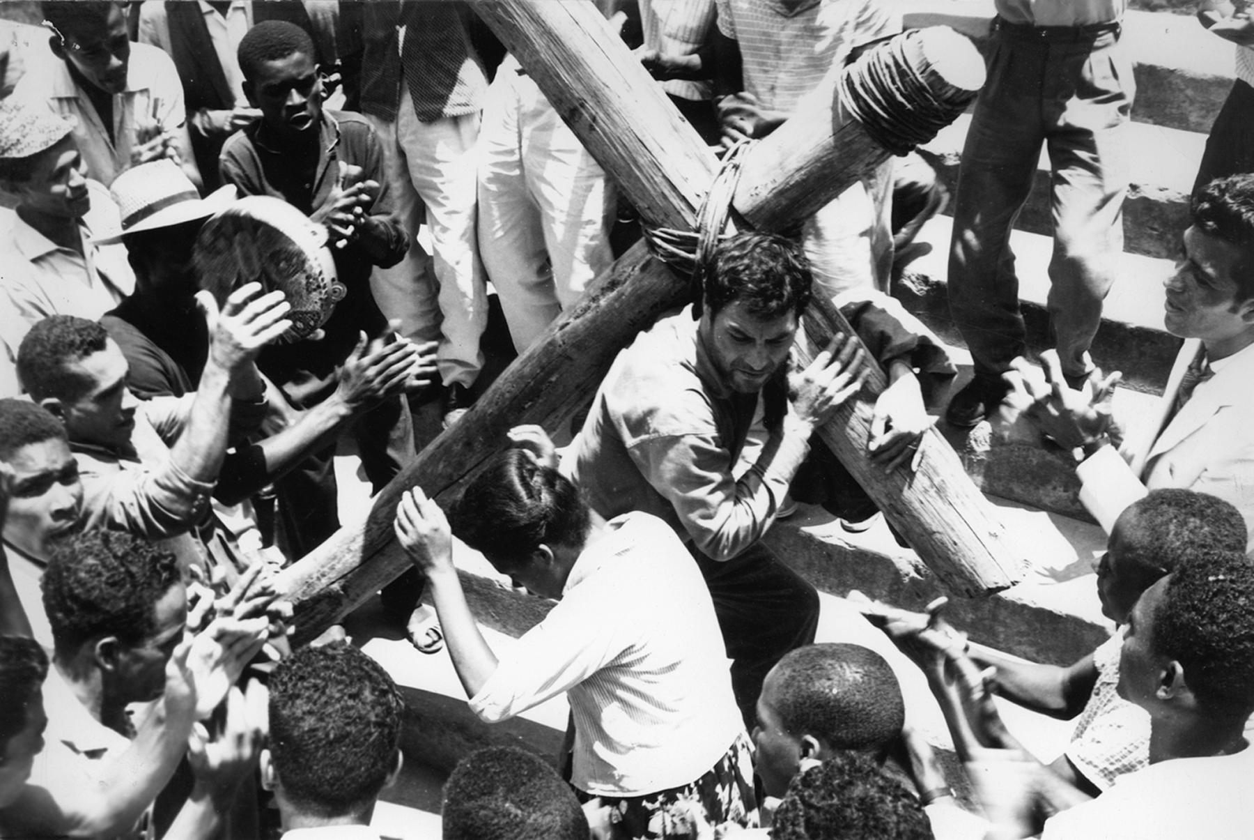 Cena de filme em preto e branco. Um homem segurando uma cruz de madeira. Ao redor, pessoas olhando para ele, e algumas pessoas batendo palmas.
