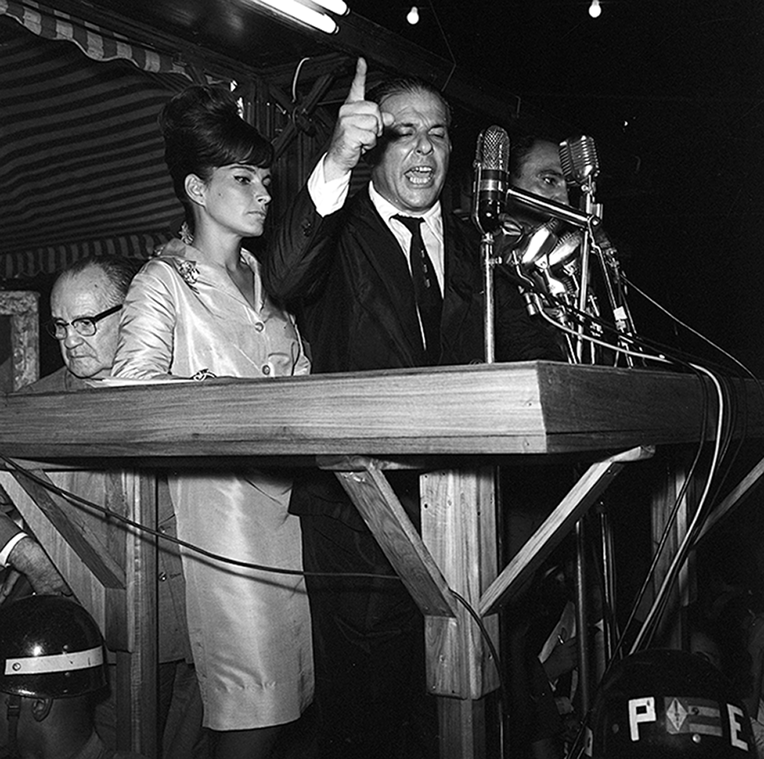 Fotografia em preto e branco. Ao centro, homem usando terno. Ele está de pé em um palanque com microfone. Ao lado, uma mulher com cabelos presos, usando um vestido.