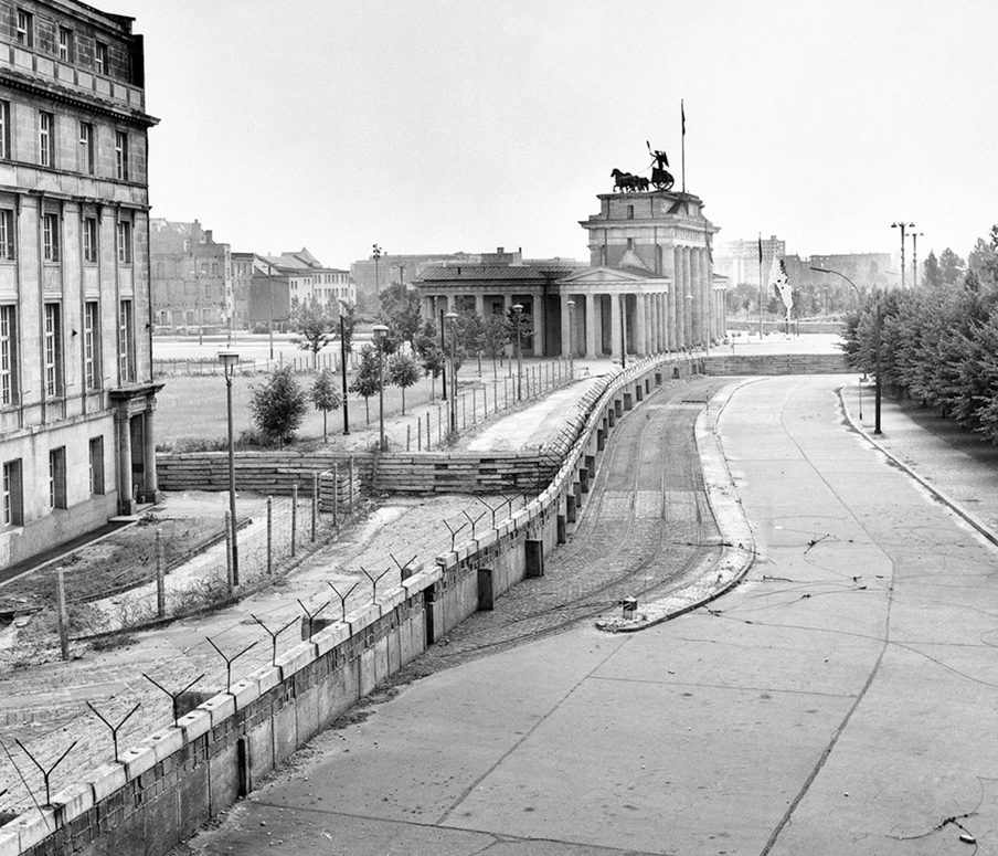 Fotografia em preto e branco. Vista de uma rua dividida por um muro alto e extenso.