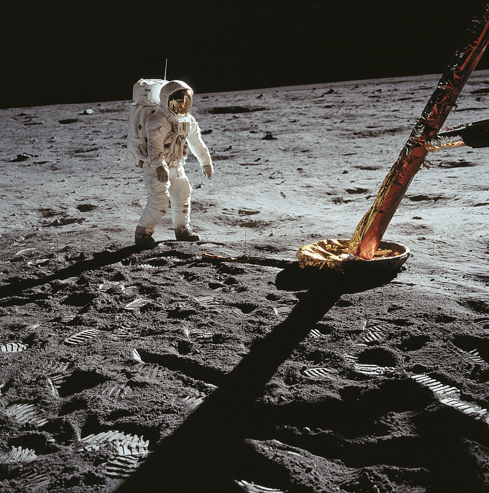 Fotografia. Uma pessoa usando roupas e capacete de astronauta caminha sobre o solo da Lua.