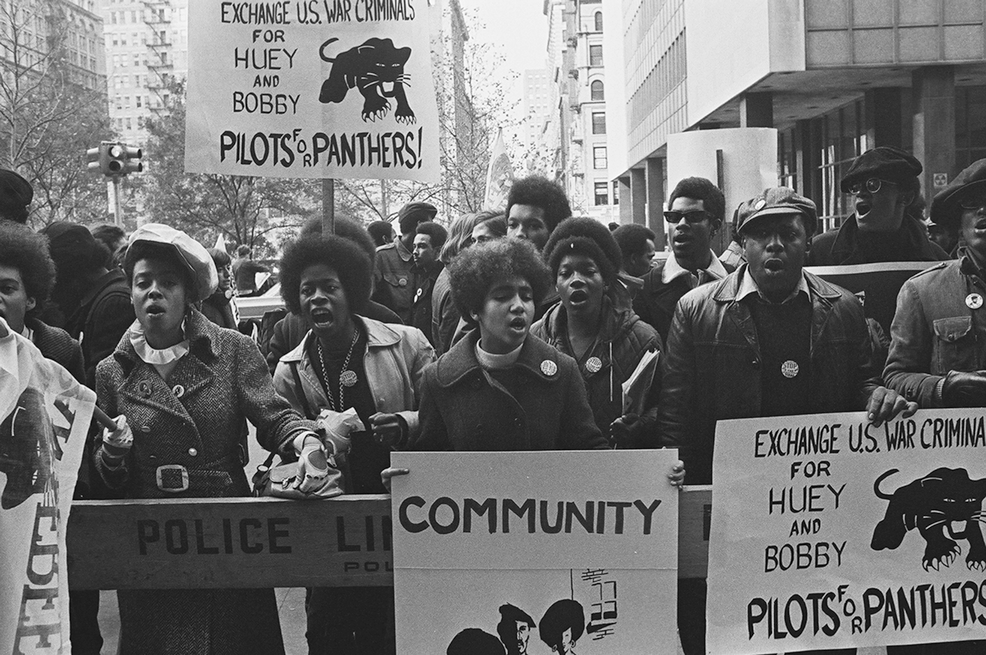 Fotografia em preto e branco. Pessoas negras em uma rua segurando cartazes.
