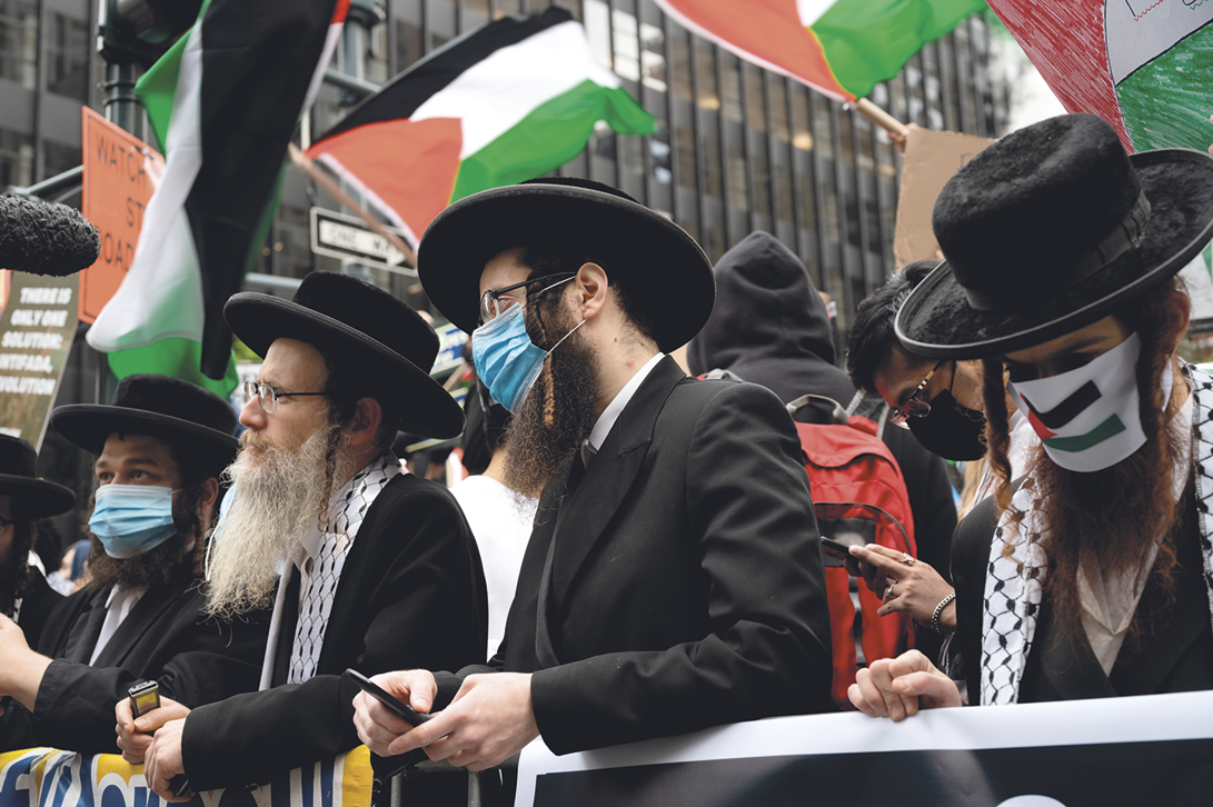 Fotografia. Homens com cabelos e barba grande, usando chapéu e terno preto. Alguns estão usando máscaras cobrindo o nariz e a boca. Acima diversas bandeiras da Palestina.