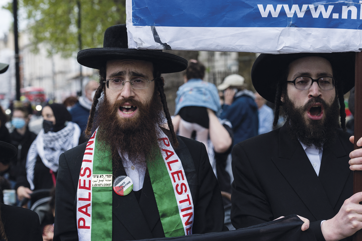 Fotografia. No primeiro plano, dois homens com barba na altura do peito, usando chapéus e camisas pretas. Um deles está com uma faixa escrito Palestine. Atrás, pessoas caminhando.