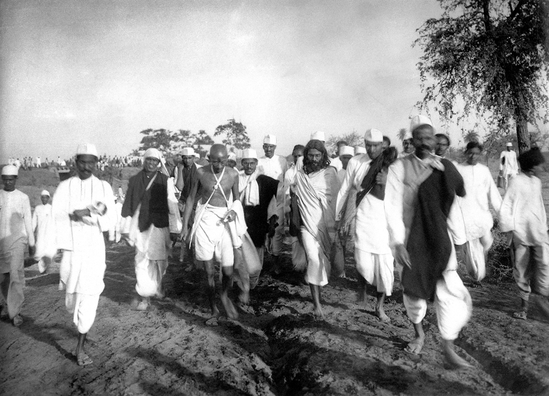 Fotografia em preto e branco. Pessoas usando roupas e chapéu branco. Eles estão descalços e caminhando em uma estrada de terra.