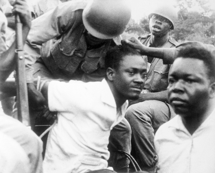Fotografia em preto e branco. Um homem negro, com camisa branca, está com os braços para trás, preso por um soldado. Atrás, outro soldado.