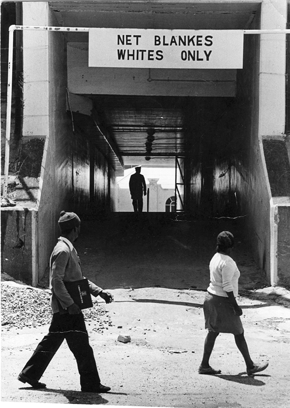 Fotografia em preto e branco. Um homem e uma mulher negra estão caminhando e olhando na direção de um túnel com a placa: NET BLANKES WHITES ONLY.