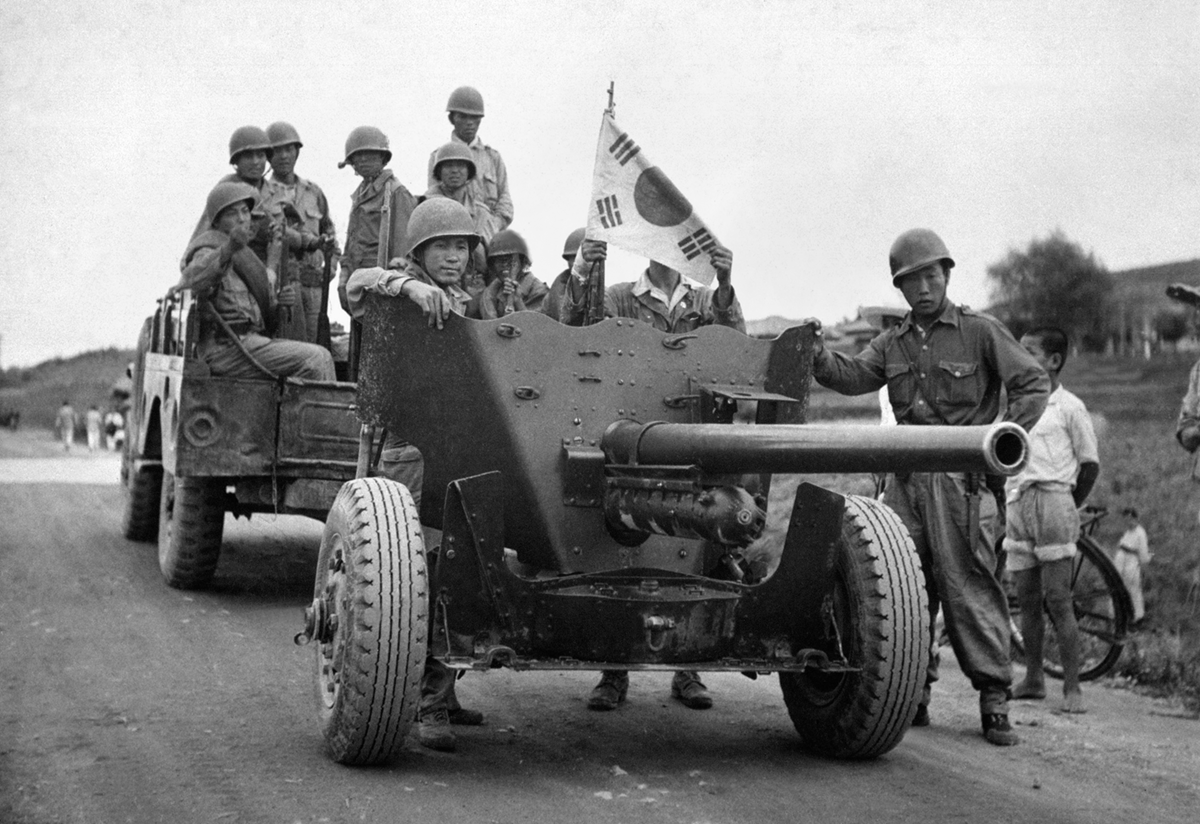 Fotografia em preto e branco. Soldados coreanos de pé em um veículo de guerra com a bandeira da Coreia do Sul hasteada.