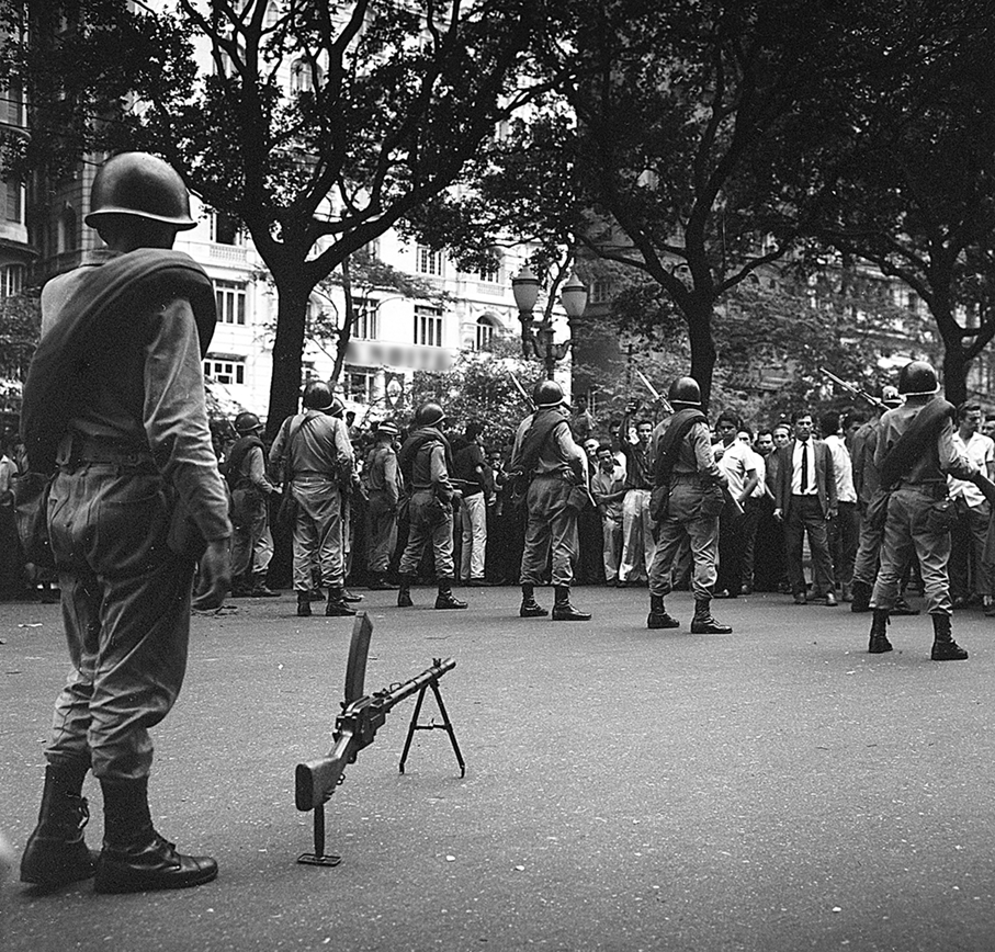Fotografia em preto e branco. No primeiro plano, à esquerda, um soldado de pé em uma rua. Ao lado, uma arma no chão. Atrás, uma fileira de soldados, seguidos de pessoas em uma rua.