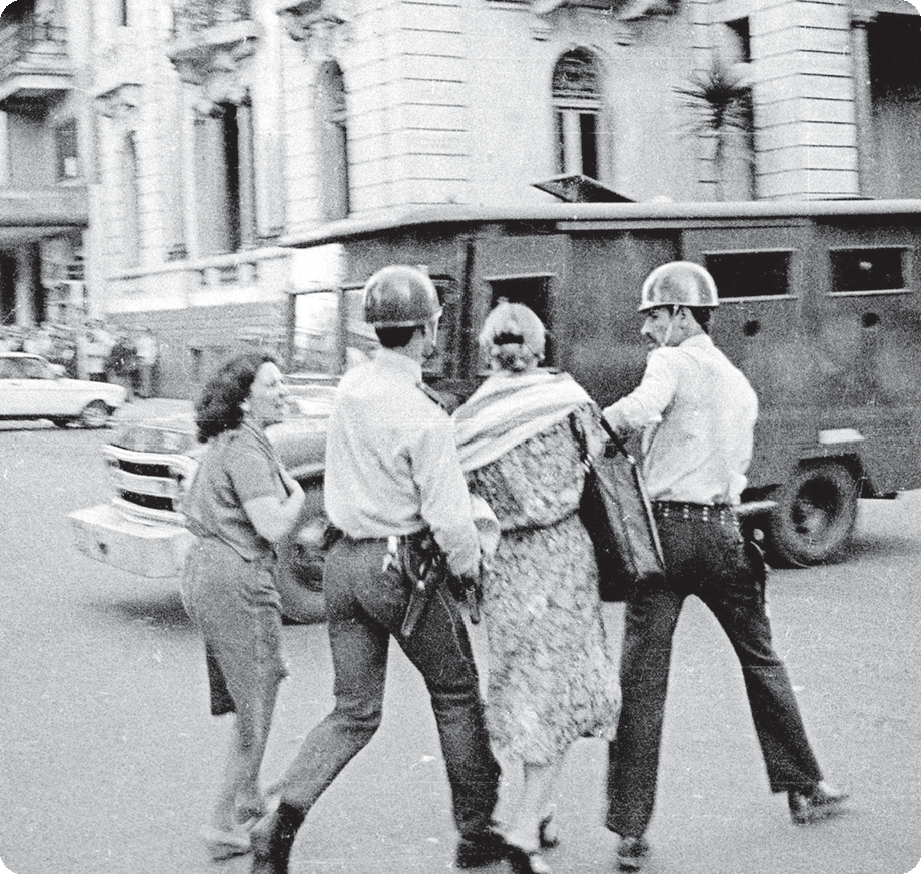 Fotografia em preto e branco. Ao centro, dois soldados segurando nos braços de uma mulher usando vestido. Ao lado, outra mulher conversa com os soldados. Ao fundo, um camburão e edifícios.