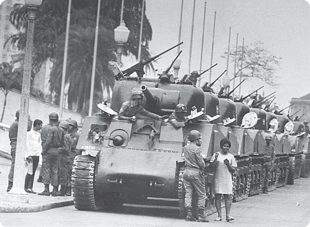 Fotografia em preto e branco. Diversos tanques de guerra enfileirados em uma rua. Ao redor, soldados e algumas pessoas.