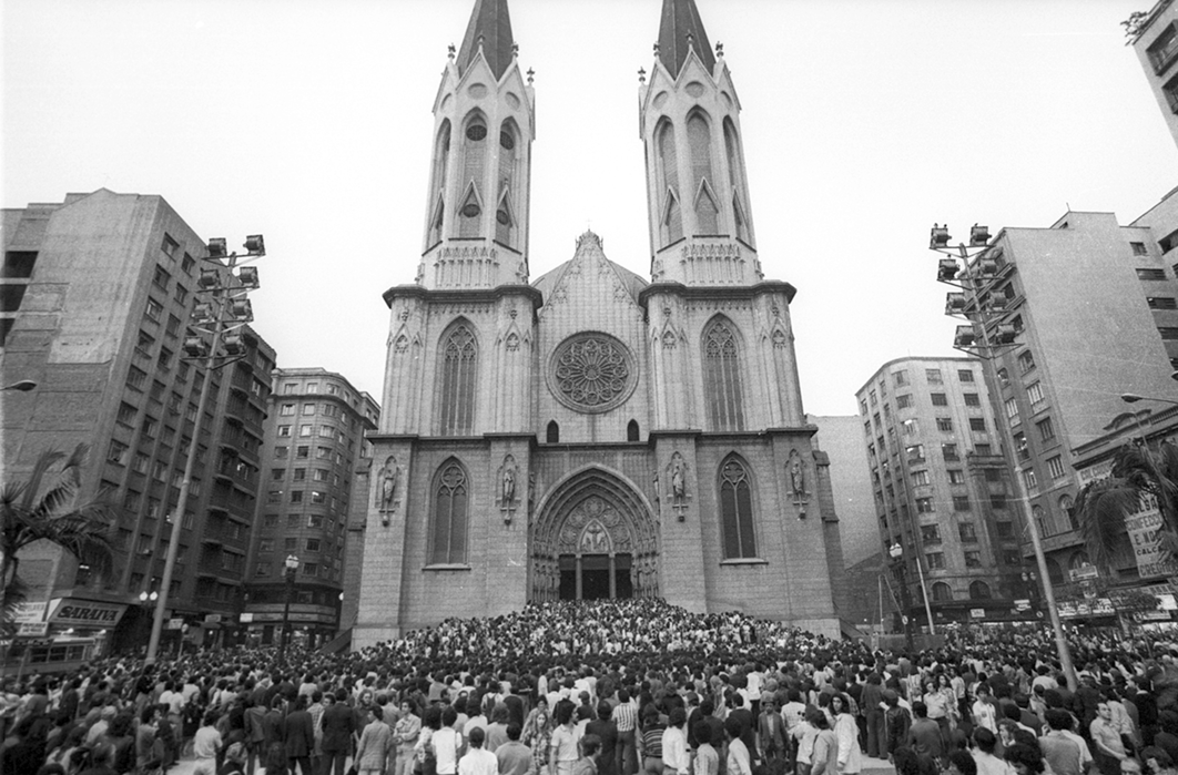 Fotografia em preto e branco. Diversas pessoas em uma rua, na frente de uma igreja com duas torres. Ao redor, diversos prédios.