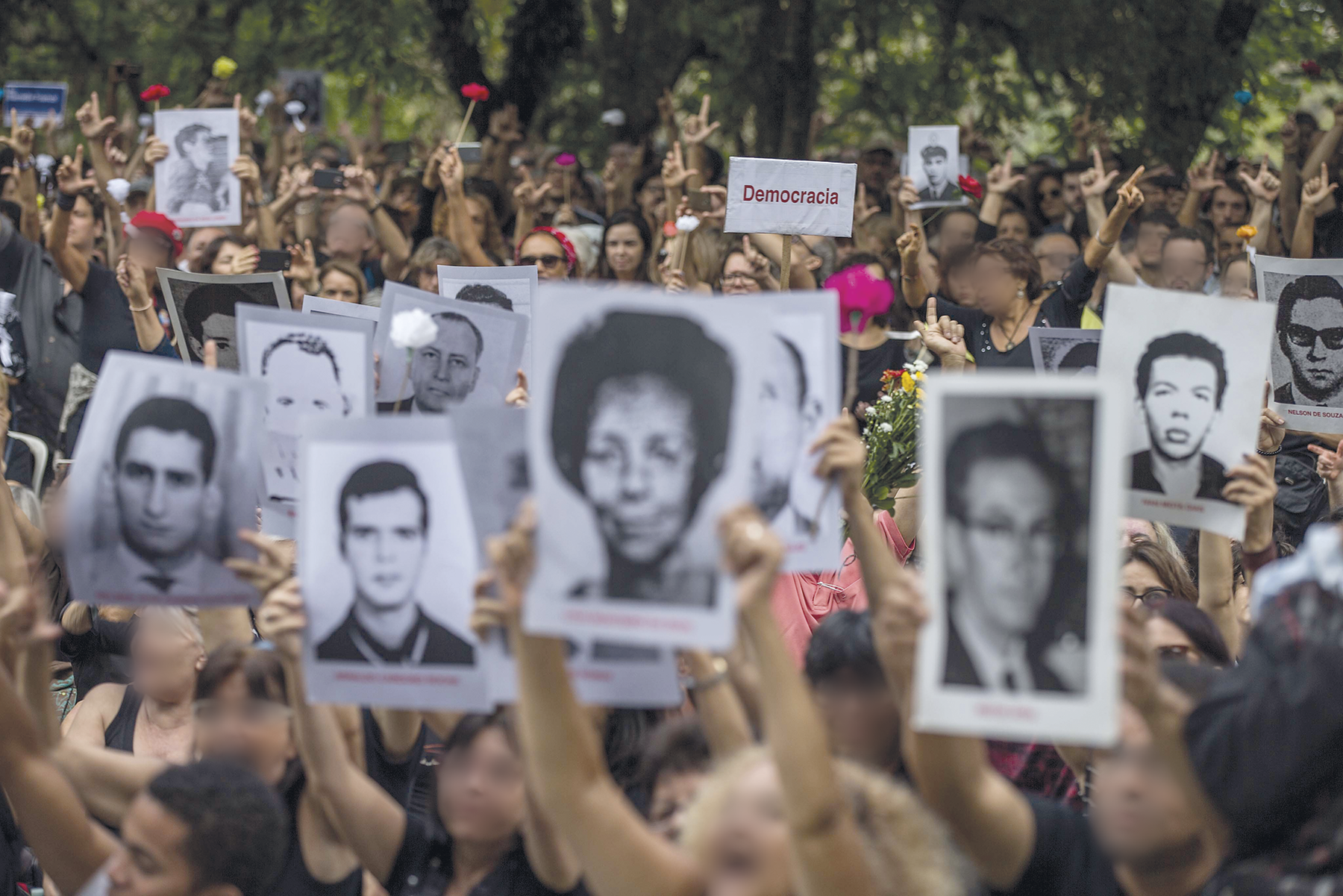 Fotografia. Destacando as mãos de diversas pessoas em uma rua segurando cartazes e fotografias em preto e branco do busto de pessoas. Entre elas, há uma placa com o texto: DEMOCRACIA.