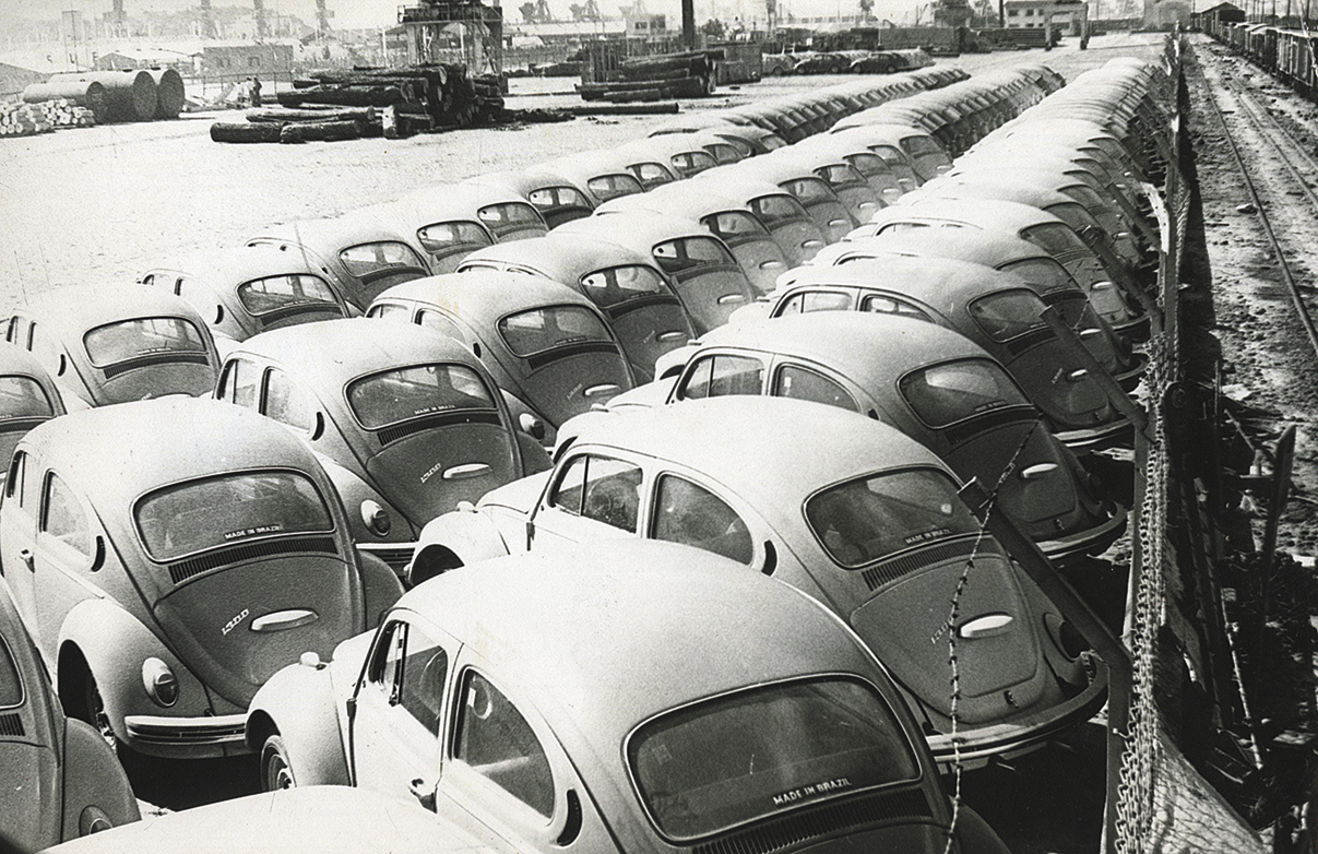 Fotografia em preto e branco. Diversos veículos da mesma marca e modelo enfileirados e um pátio, um do lado do outro.