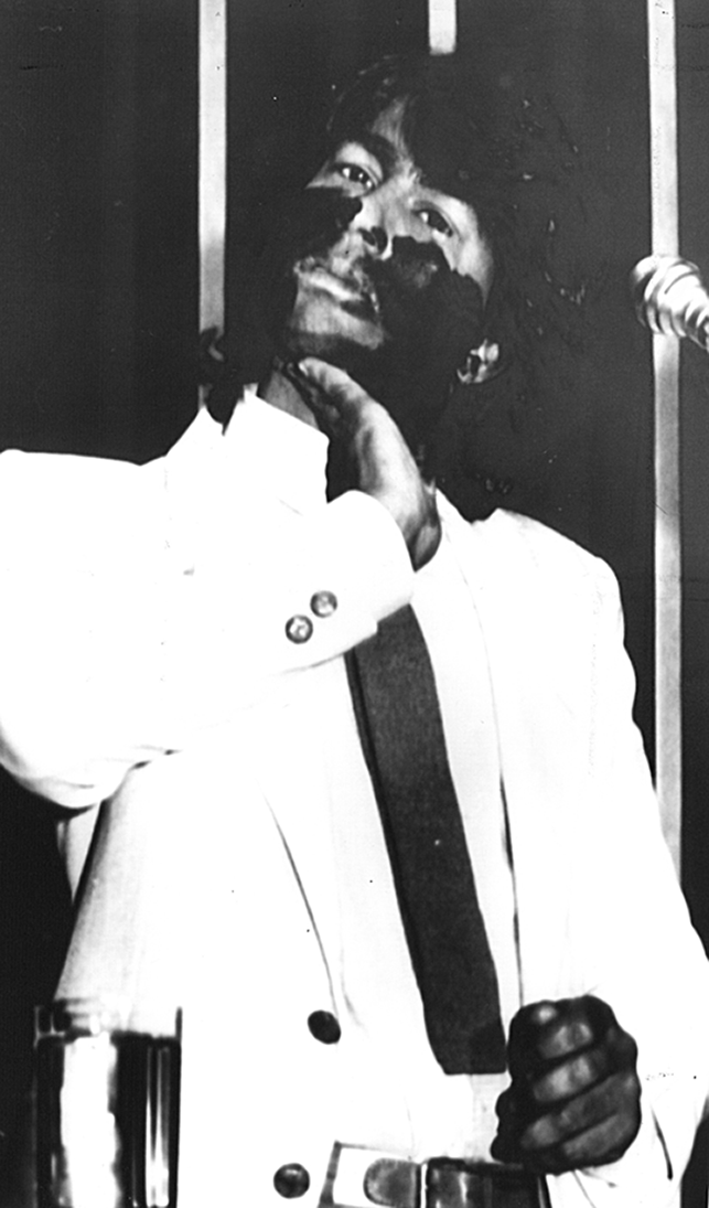Fotografia em preto e branco. Destacando o busto de um homem com cabelos liso, usando terno branco. Ele está com a mão sobre o rosto pintado de tinta.