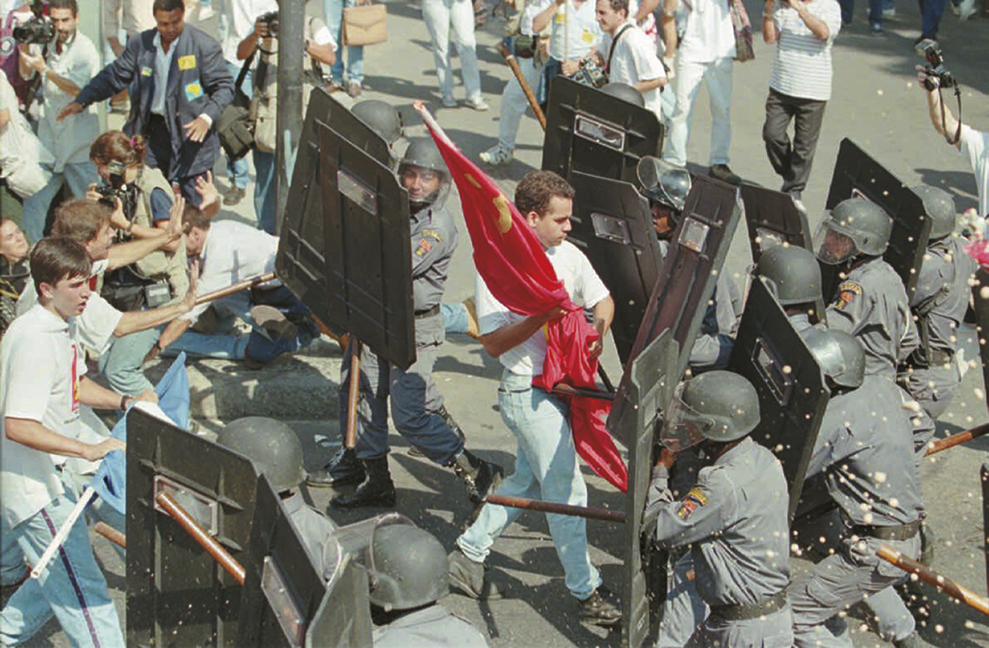 Fotografia. À esquerda jovens uniformizados segurando bandeiras. À direita, policiais segurando bastões e escudos. Entre eles, há um jovem caminhando com uma bandeira vermelha entre os policiais.