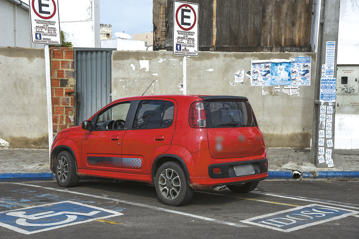 Fotografia. Um carro vermelho estacionado em uma vaga. No chão, o texto: IDOSO.