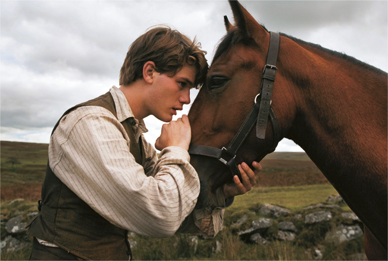Cena de filme. Um jovem com cabelos loiros, usando camisa e colete. Ele está de perfil com as mãos e a cabeça encostada no focinho de um cavalo.