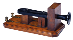 Fotografia de um antigo telefone, com estrutura de madeira e parte metálica semelhante a parte de um instrumento musical, trompete.