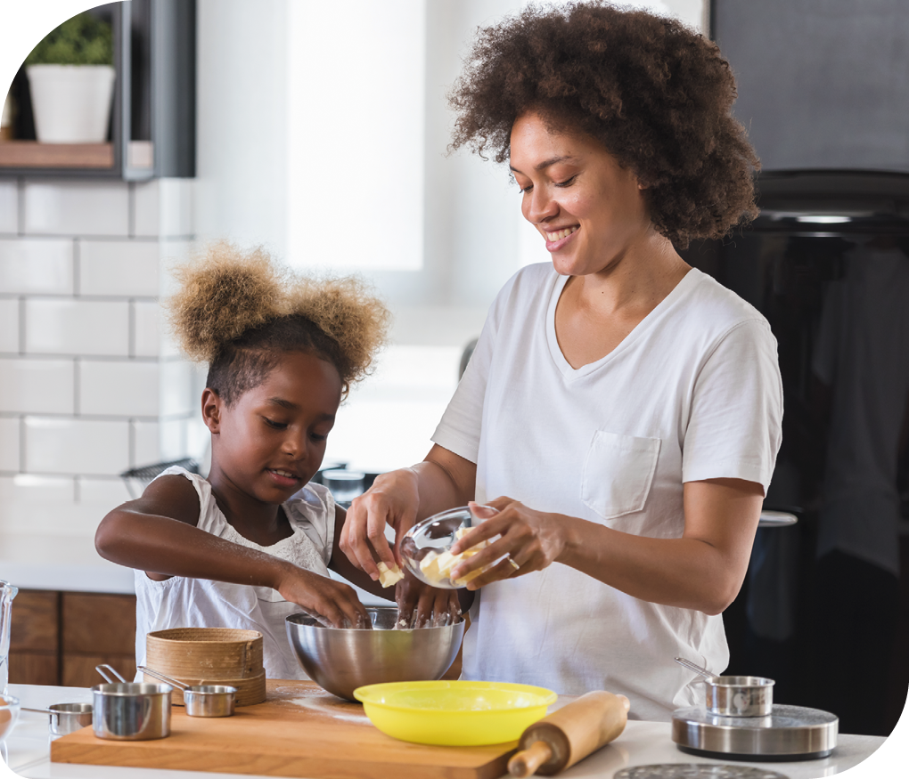 Fotografia de uma mulher e uma menina diante de uma mesa, cozinhando. A mulher está segurando um pequeno pote com um ingrediente e a menina está coma mãos dentro de um recipiente. Sobre a mesa há uma tábua, um rolo de massa e outros potes.